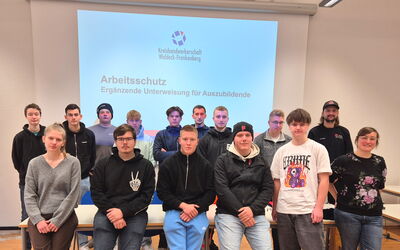 Azubis sitzen und stehen vor einer PowerPoint-Folie. Rechts daneben steht Dozentin Eva Maria Hartmann.