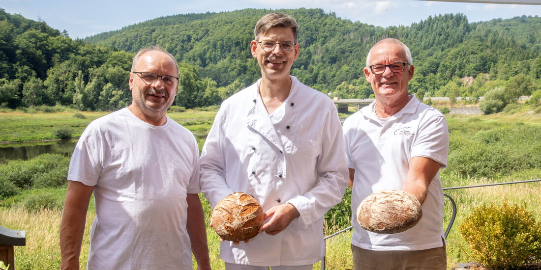 Drei Männer stehen auf der Terrasse der Bäckerei Raabe mit leckerem Brot in der Hand  – darunter der Obermeisterder, Beisitzer der Innung und Prüfer.