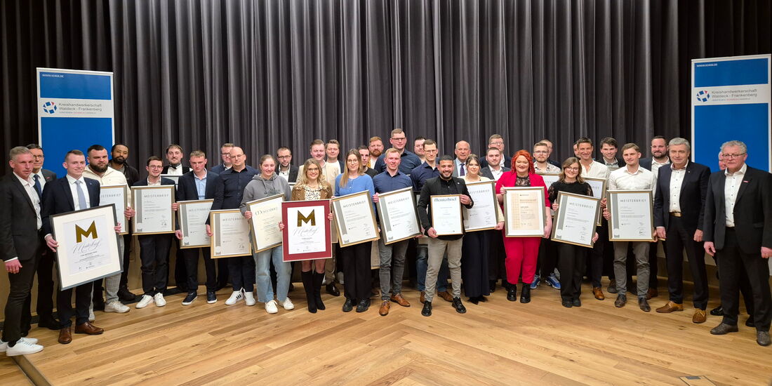 Das Foto zeigt eine große Gruppe frischgebackener Meisterinnen und Meister, die stolz mit ihren Meisterurkunden in der Hand vor der Bühne im Philipp-Soldan-Forum in Frankenberg stehen. Sie tragen festliche Kleidung, einige in Anzügen.