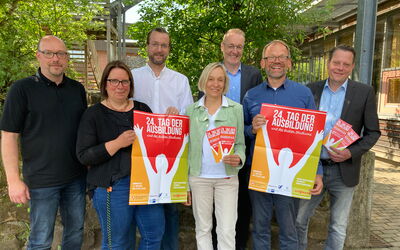 Sieben Veranstalter*innen mit Messeplakat und Flyern für den Tag der Ausbildung und des dualen Studiums vor der Ortenbergschule in Frankenberg.