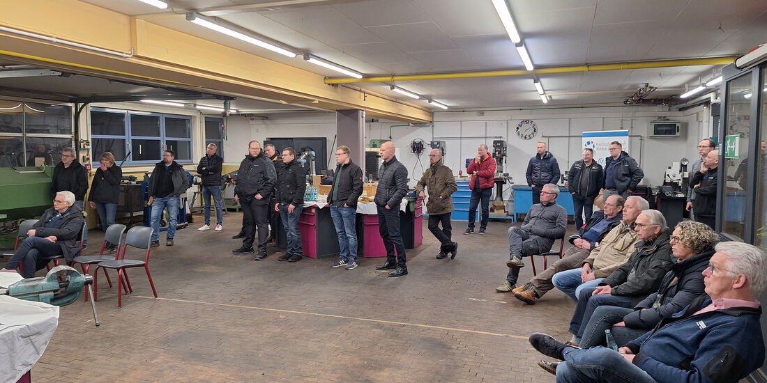 Das Foto zeigt eine Gruppe von Menschen in einer Schweiß-Werkstatt. Die Teilnehmenden stehen oder sitzen in einem Halbkreis und hören einem Vortragenden zu. Sie tragen Alltagskleidung. Viele der Zuhörer blicken gespannt auf den Vortragenden.