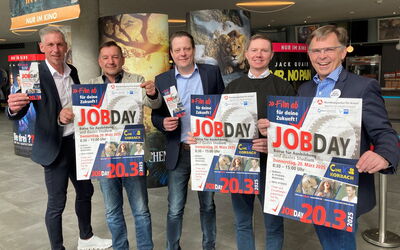 Fünf Männer stehen gemeinsam im Foyer des Korbacher Kinos und blicken lächelnd in die Kamera. Sie halten Plakate und Flyer zum Jobday 2025 in den Händen, um auf die Veranstaltung aufmerksam zu machen.